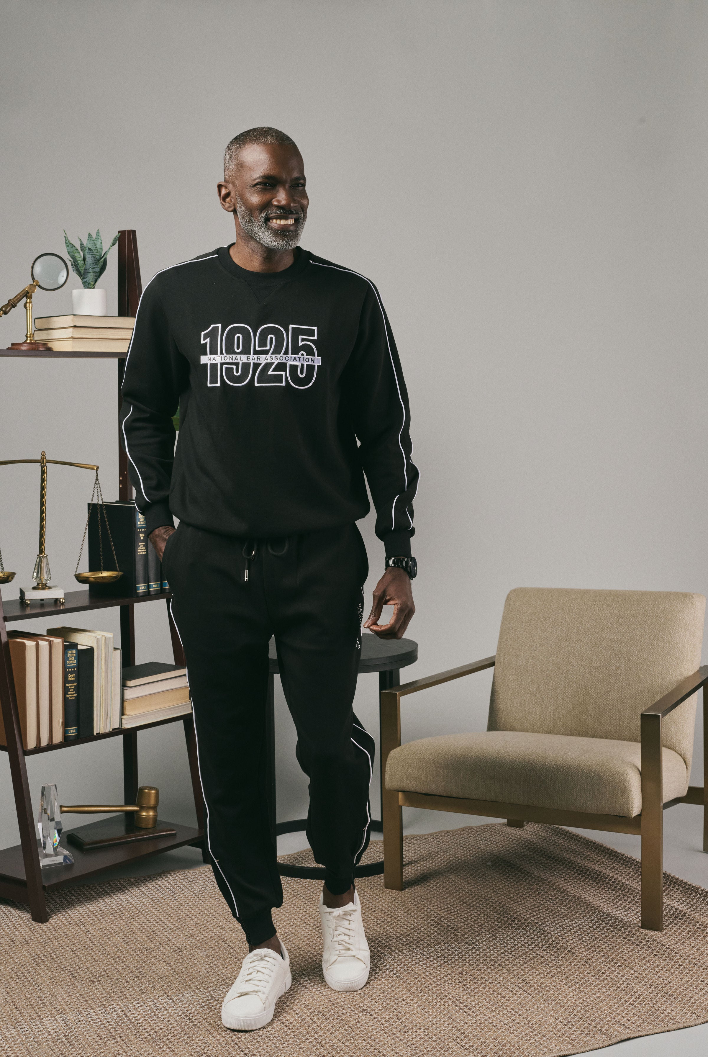 Man wearing a black sweatshirt and black joggers with white text in a modern room with a chair and a bookshelf