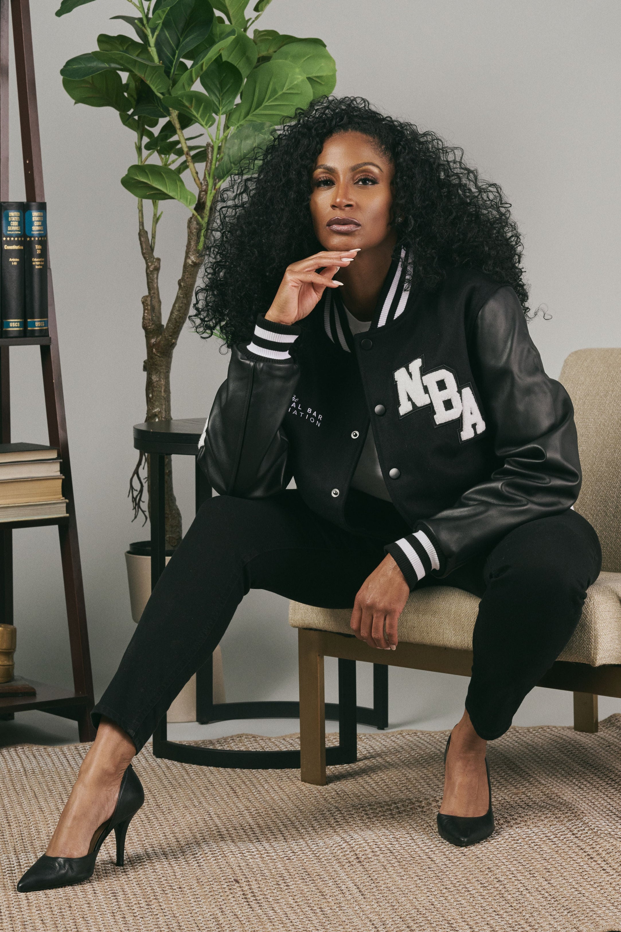 Woman wearing a NBA varsity jacket sitting on a chair in a room with books and plants.