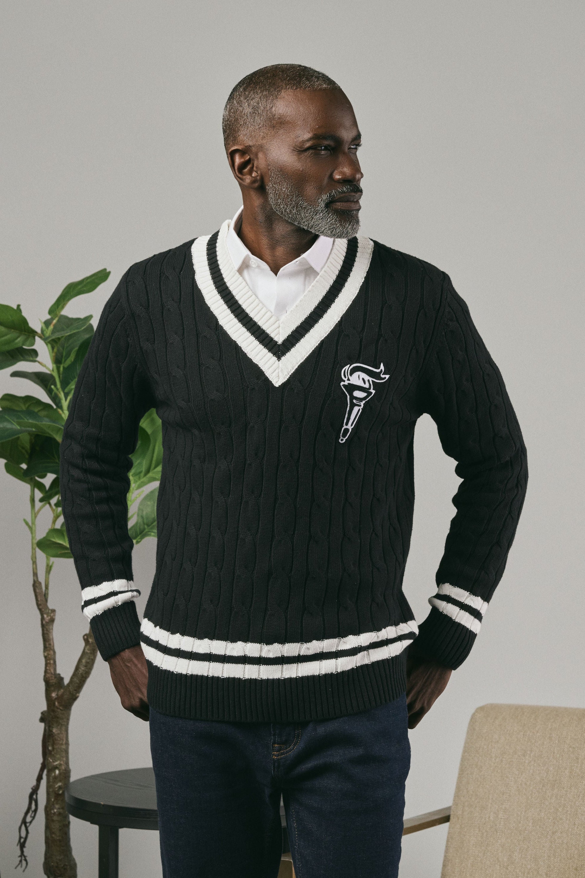 Man wearing a black sweater with a logo, standing indoors and looking sideways against a neutral background.