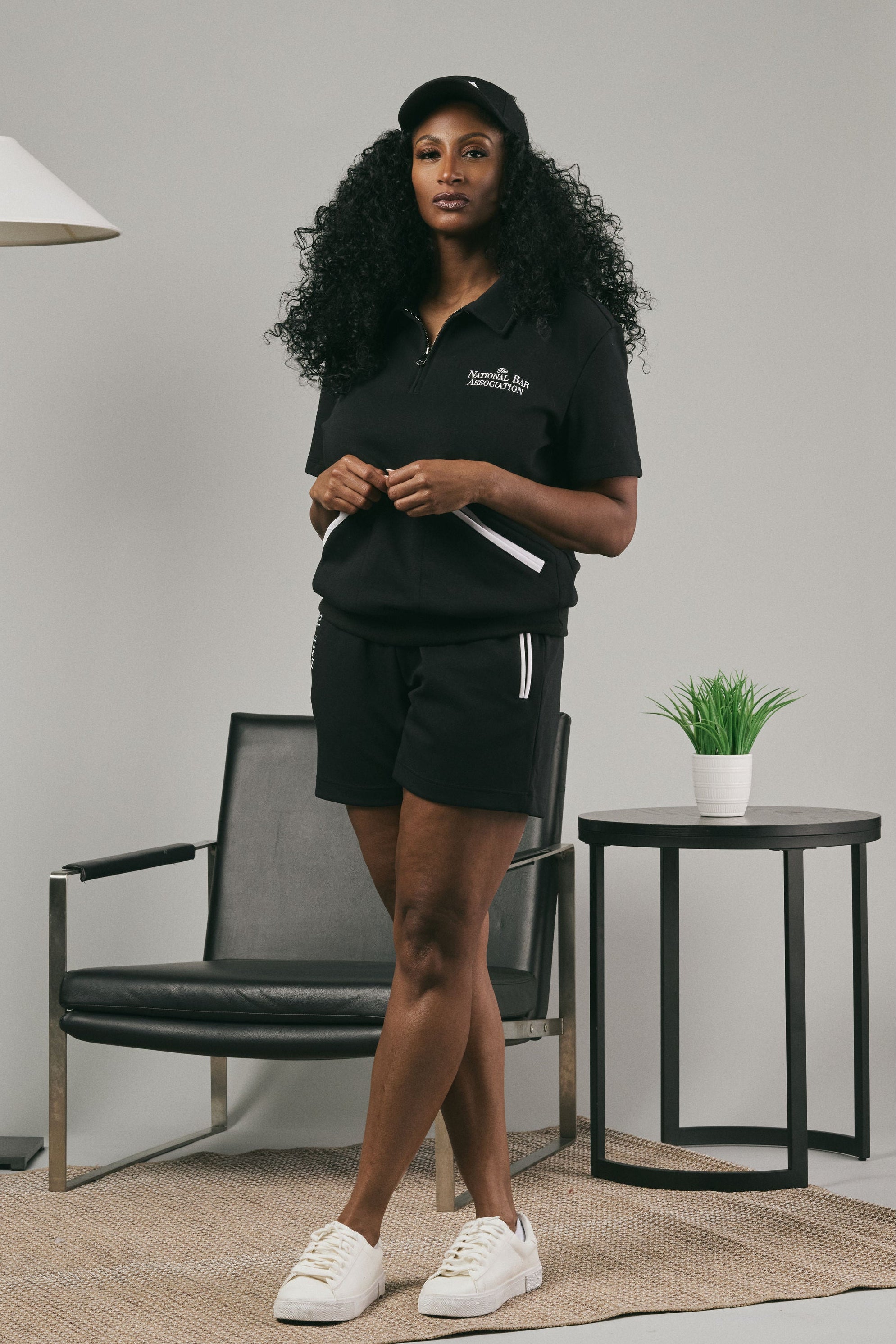Woman wearing a black pullover and black shorts standing in a modern room with a chair and lamp.