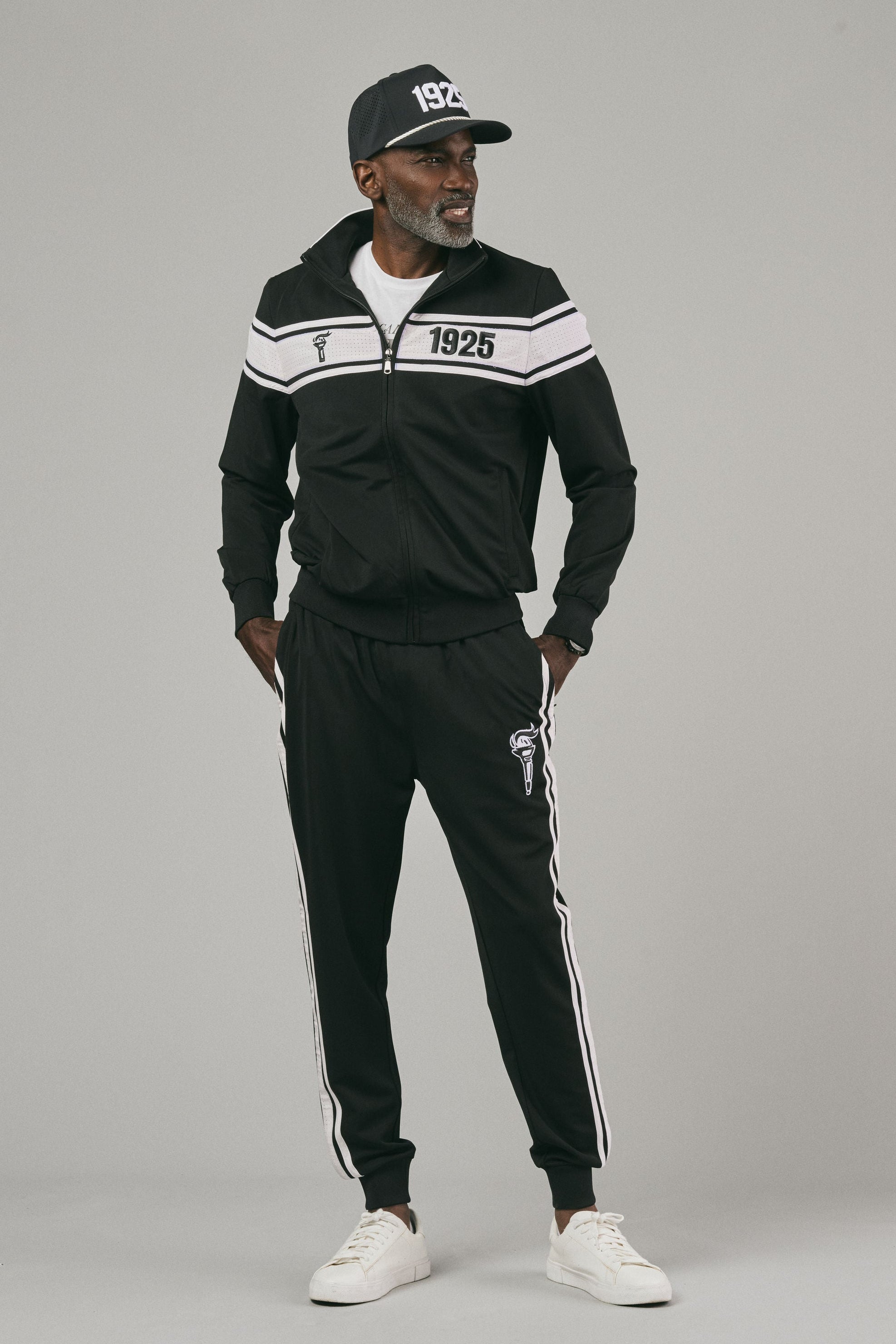Man wearing a black tracksuit with white stripes and logo and a cap looking sideways a gray background