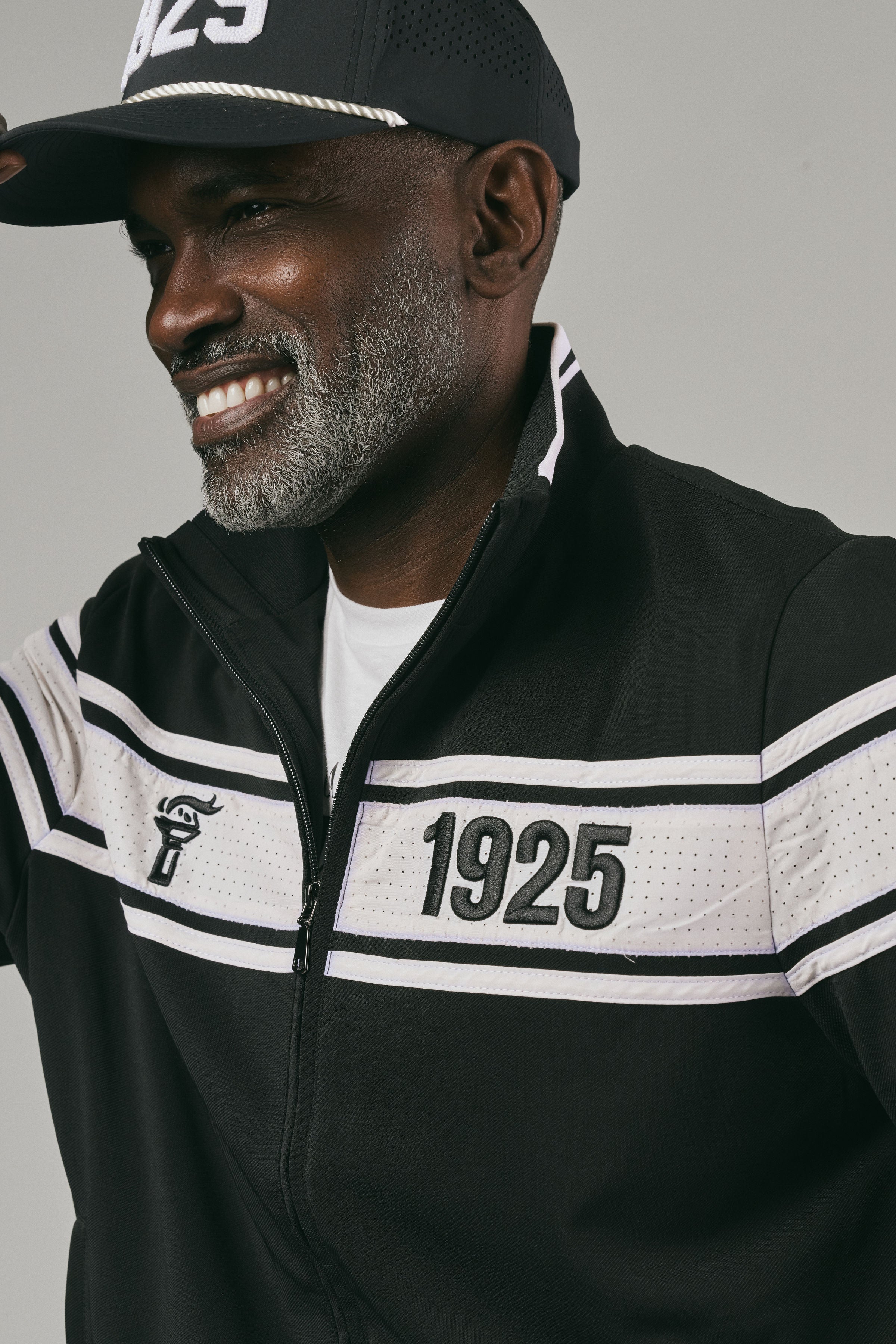 Close up of a man wearing a black and white jacket with '1925' and a logo on a grey background
