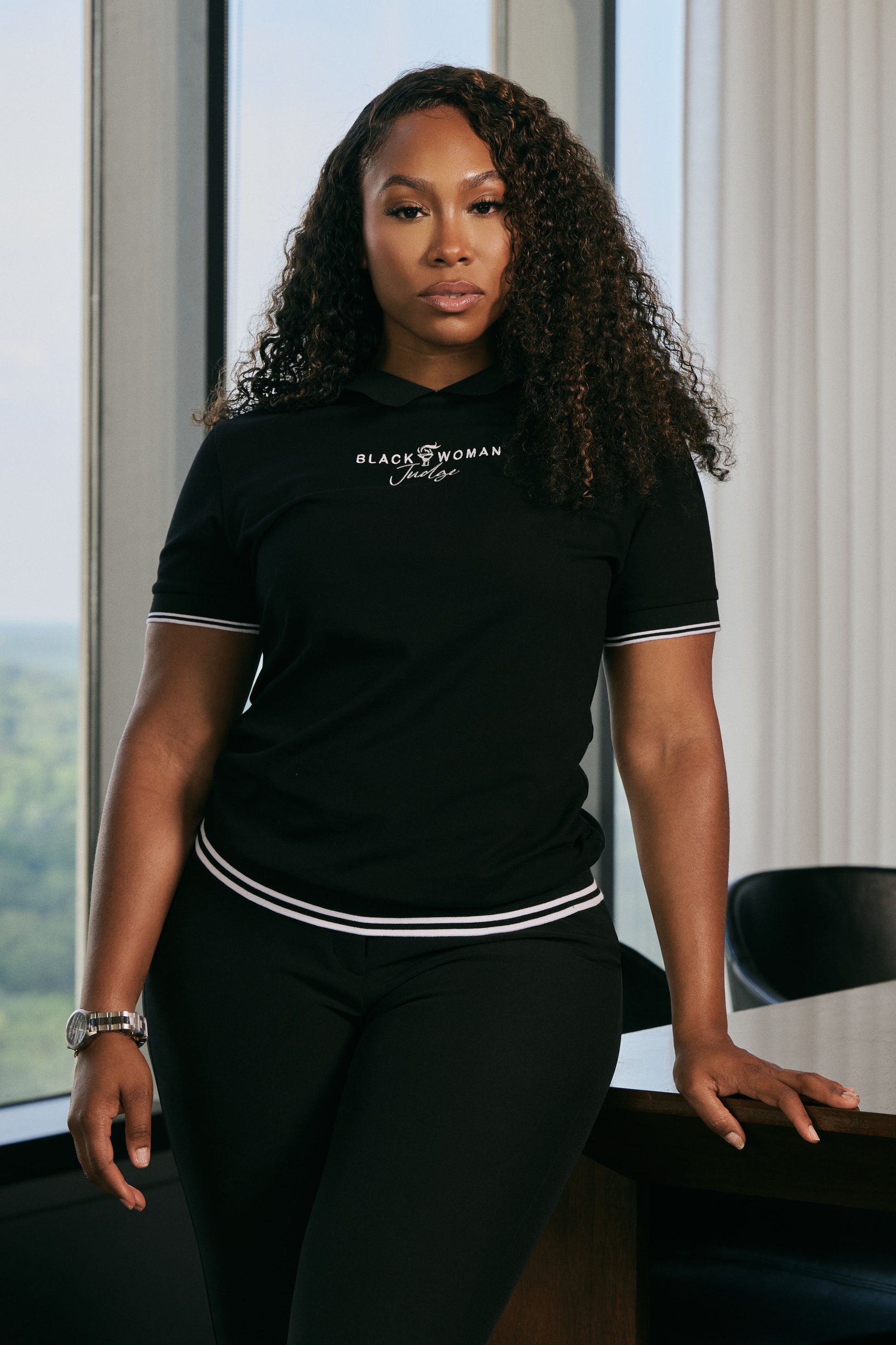 Woman wearing a black t-shirt with text, standing in an office setting with large windows.
