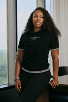 Woman wearing a black t-shirt with text, standing by a window with a view of greenery.