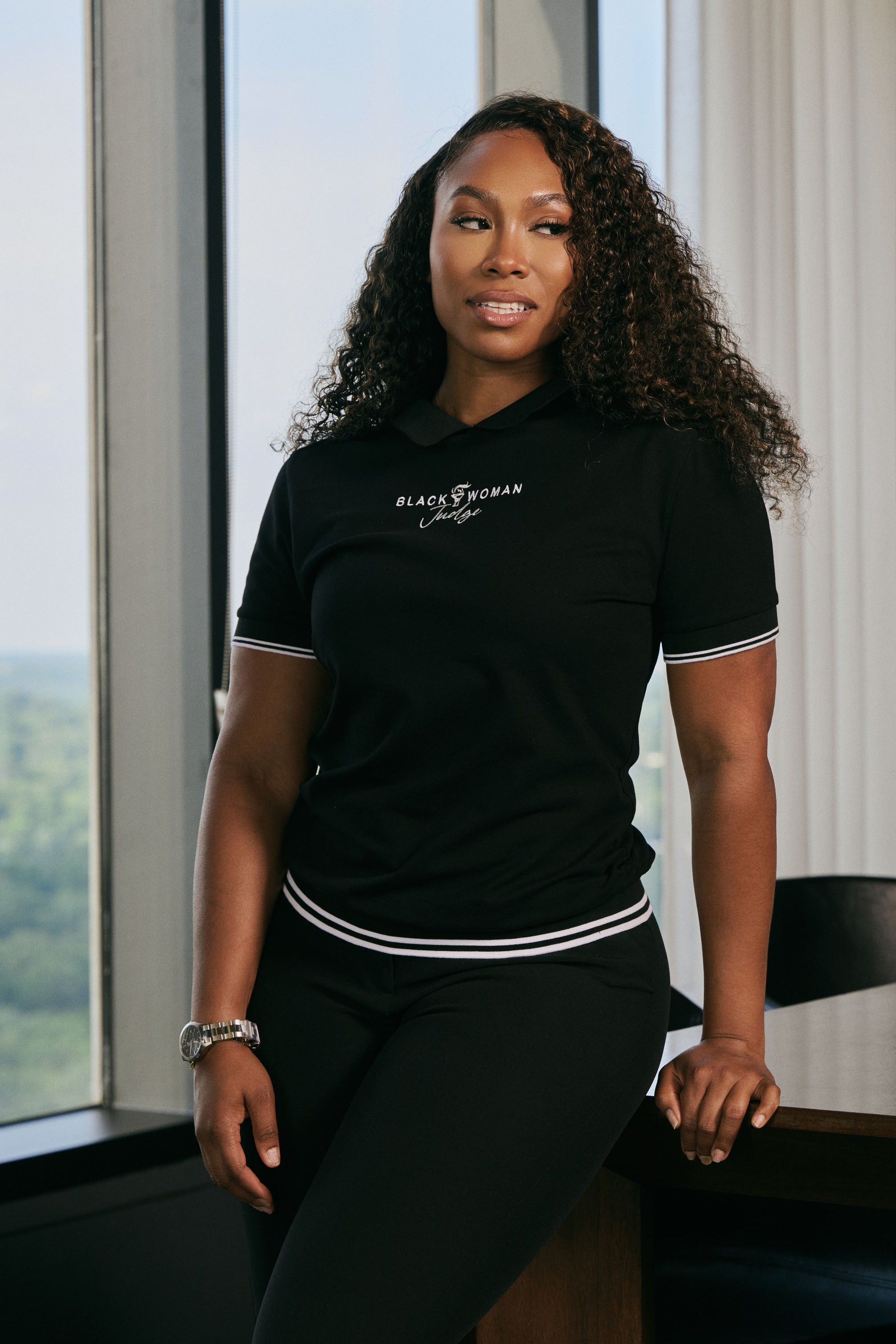 Woman wearing a black t-shirt with text, standing by a window with a view of greenery.