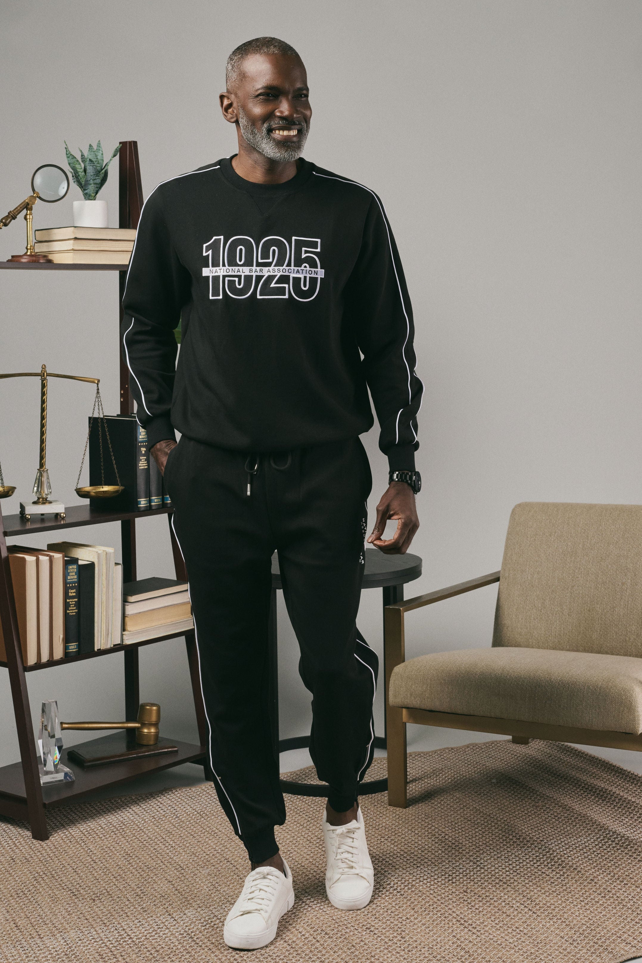 Man wearing a black sweatshirt with '1925' on it in a room with a chair and bookshelf.