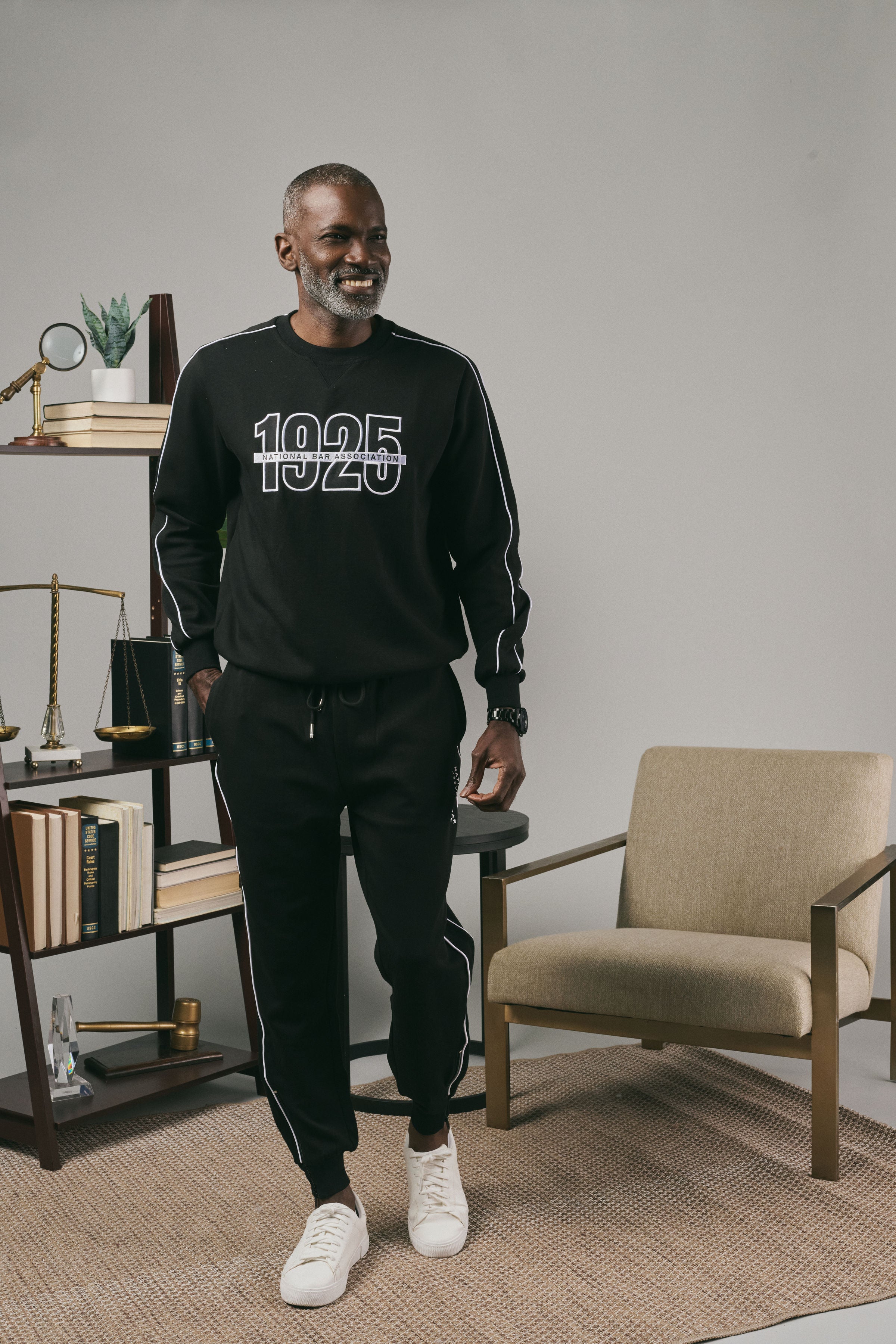 Man wearing a black sweatshirt and black joggers with white text in a modern room with a chair and a bookshelf