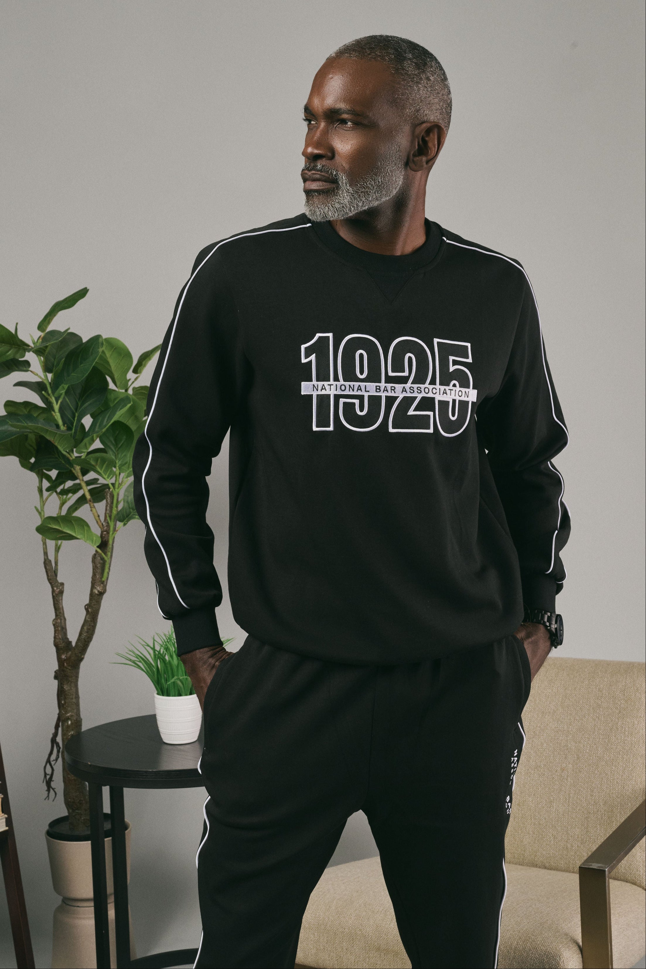 Man wearing a black crewneck with '1025' printed on it, standing in a room with a plant and furniture.