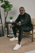 Man sitting on a chair wearing a black tracksuit with white accents, in a room with a plant and small table.