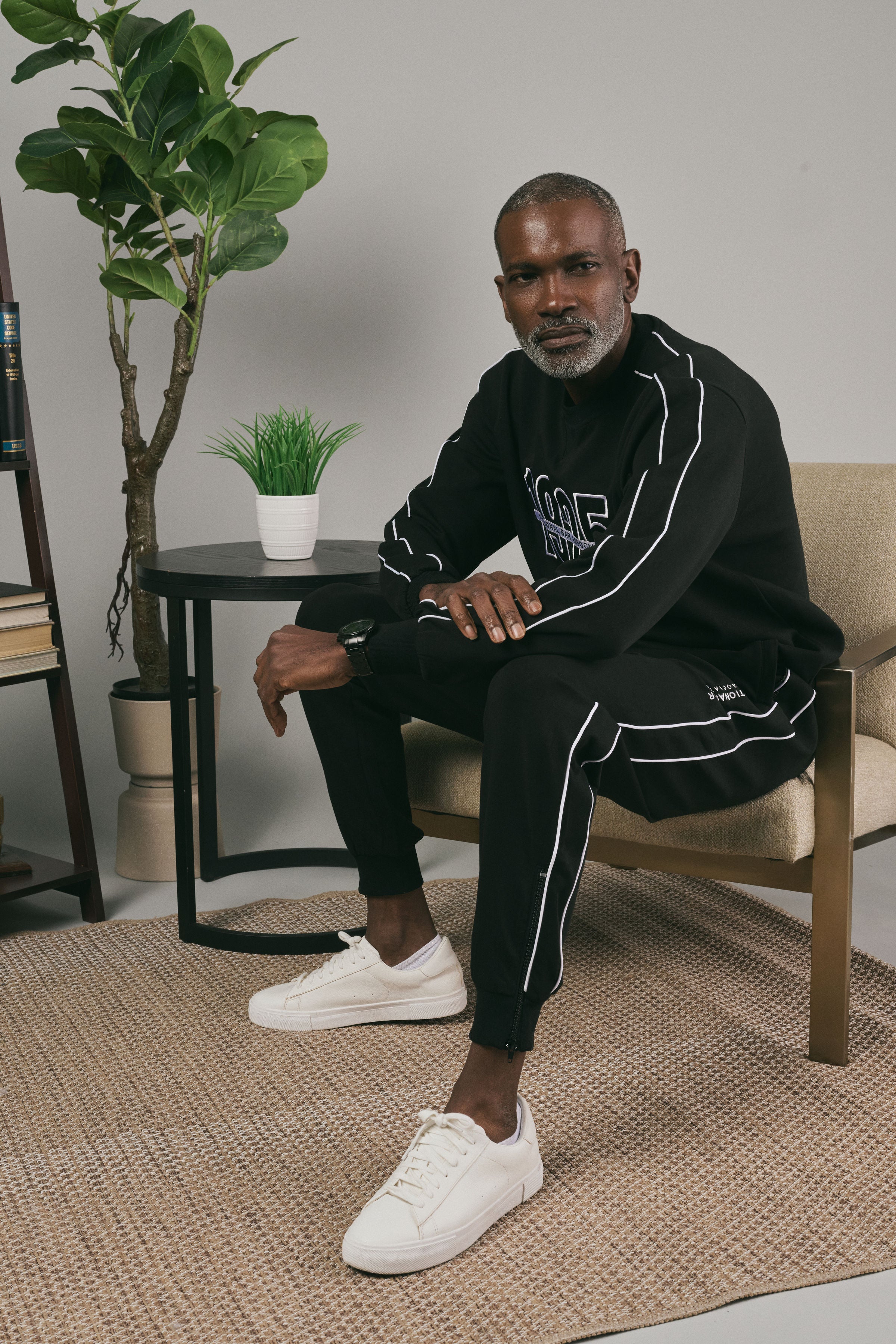 Man sitting on a chair wearing a black tracksuit with white accents, in a room with a plant and small table.