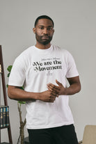 Man wearing a white t-shirt with 'We are the Movement' text, standing indoors.