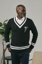 Man wearing a black sweater with a logo, standing indoors and looking sideways against a neutral background.