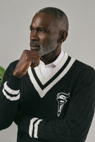 Man wearing a black sweater with white accents and a logo, posing against a grey background