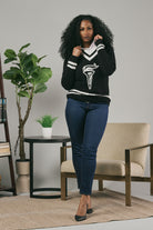 Woman wearing a black sweater with a white design in a room with a chair, table, and plants.