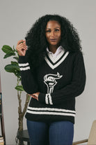 Woman wearing a black sweater with a logo in a room with books and a plant.