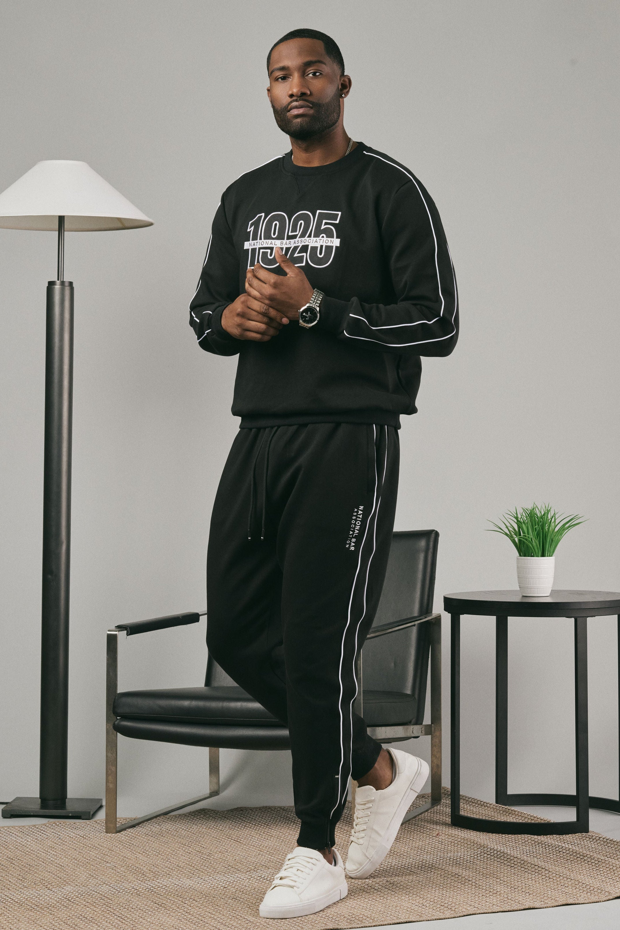 Man wearing a black sweatshirt and black joggers with white text in a modern room.