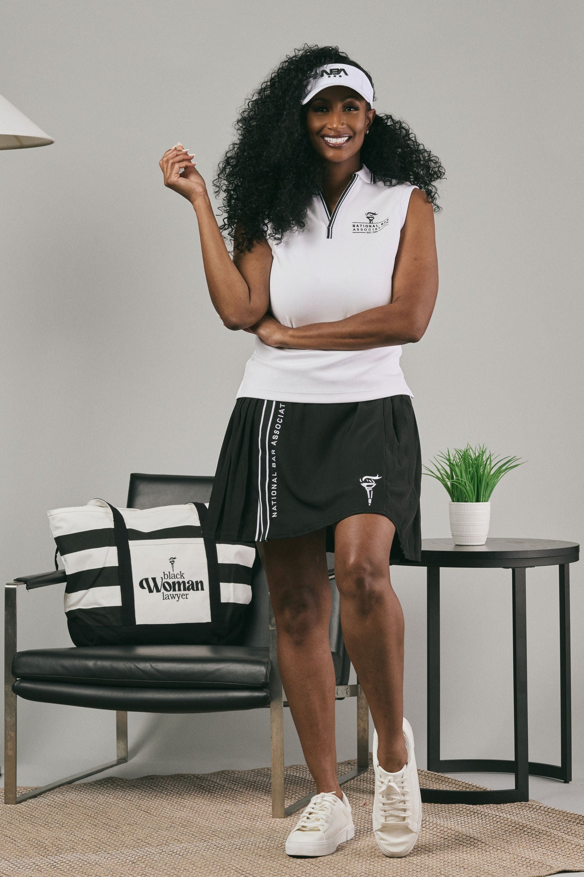 Woman in white top and black pleated skirt standing next to a chair with a bag on it, against a plain background