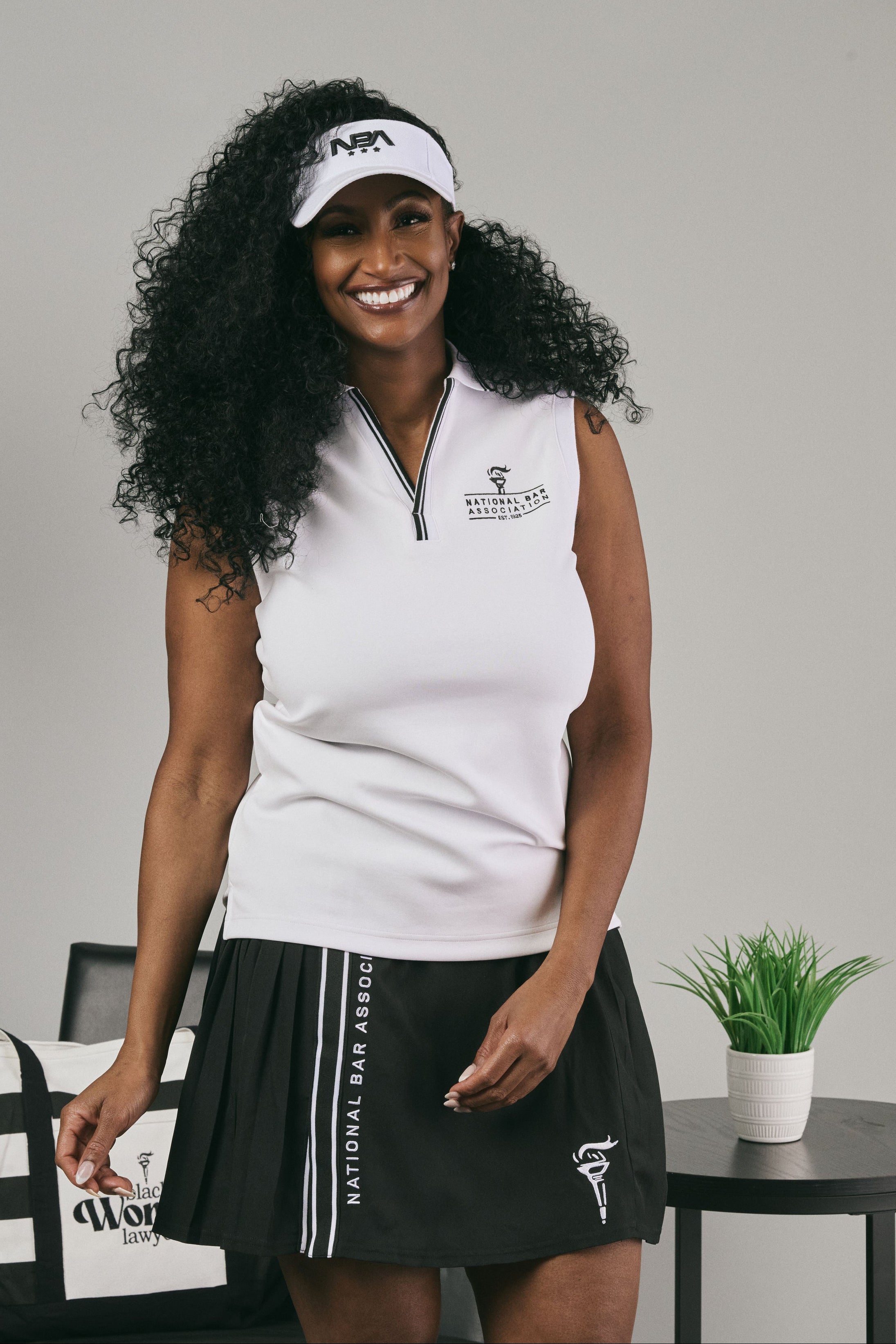 Woman wearing a white sleeveless top and black pleated skirt with a logo, standing indoors.