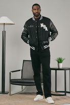 Man wearing a black varsity jacket with 'NBA' branding in a modern interior setting.