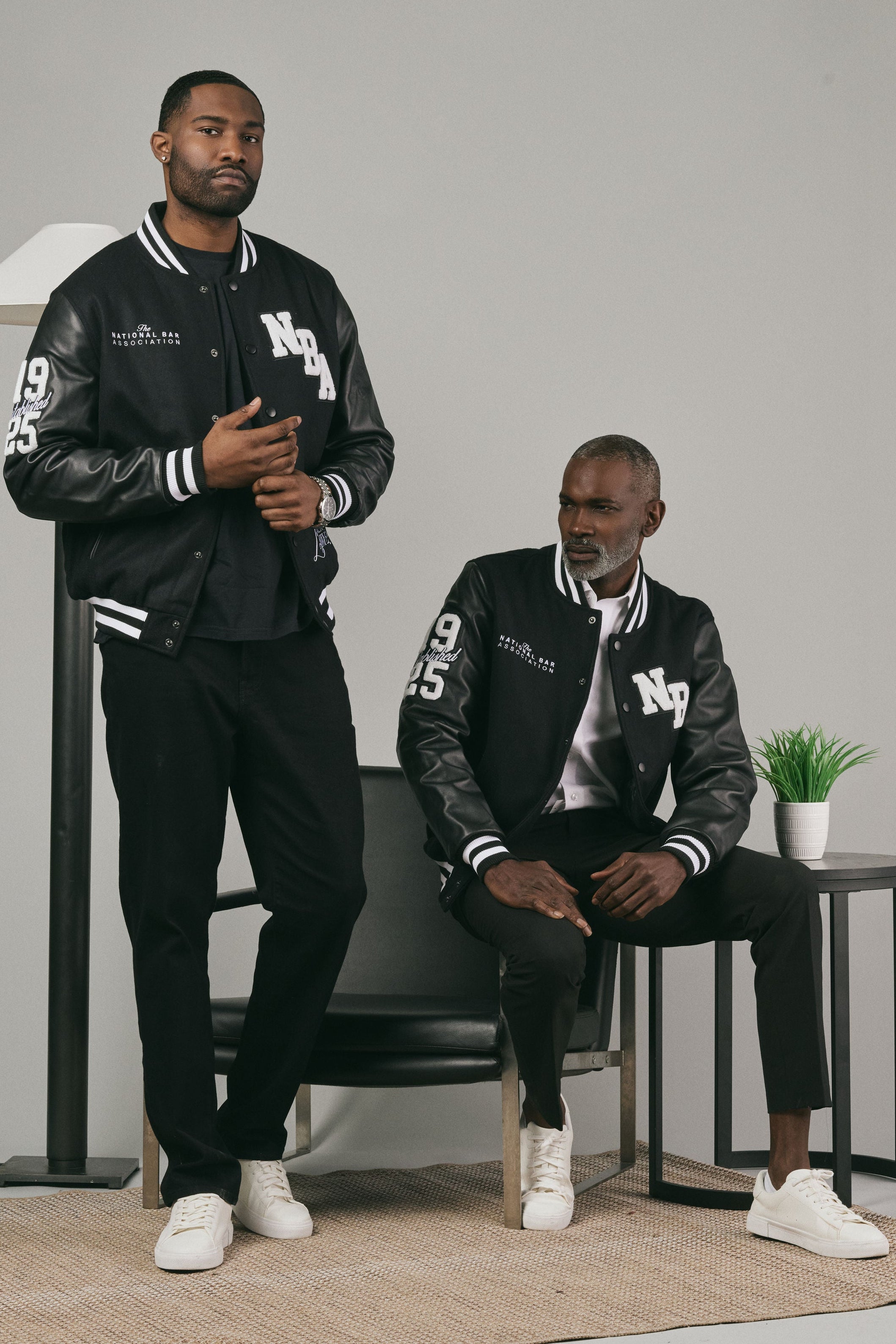 Two men wearing matching black and gray varsity jackets with white accents in a minimalistic setting.
