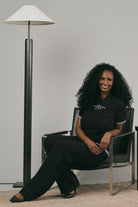 Woman wearing a black polo shirt with text and black pants, sitting in a chair in a modern interior setting with a lamp and plant.