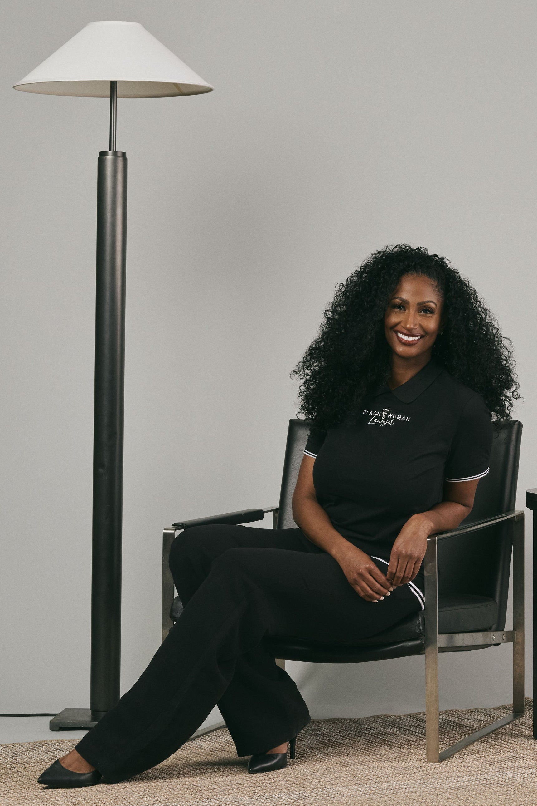 Woman wearing a black polo shirt with text and black pants, sitting in a chair in a modern interior setting with a lamp and plant.