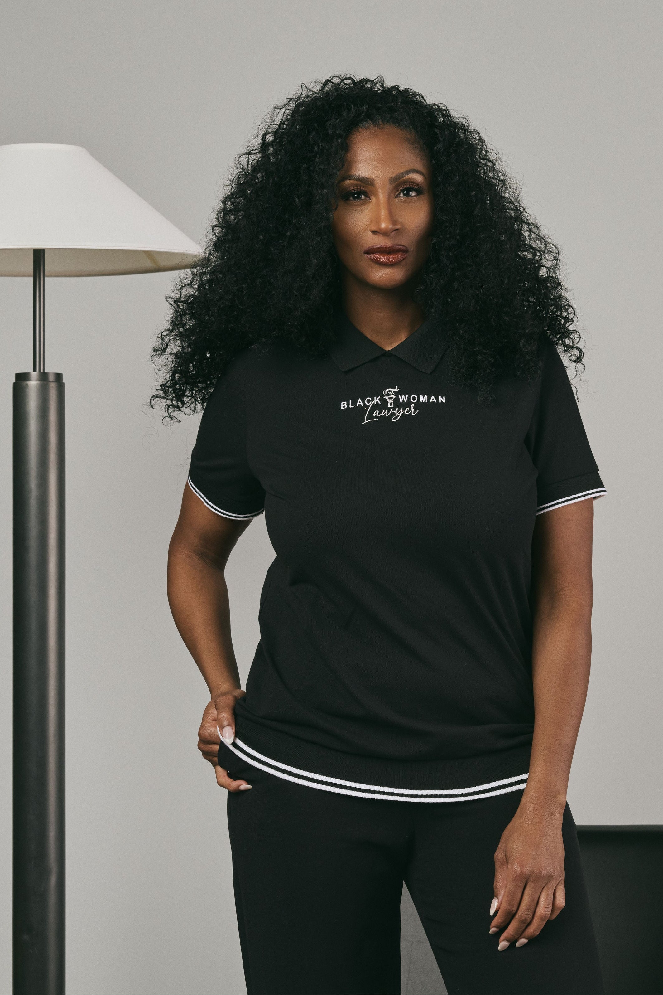 Woman wearing a black t-shirt with text 'Black Woman Lawyer', standing in a room with a lamp and chair.