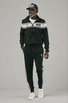 Man wearing a black tracksuit with white stripes and a cap posing against a grey background