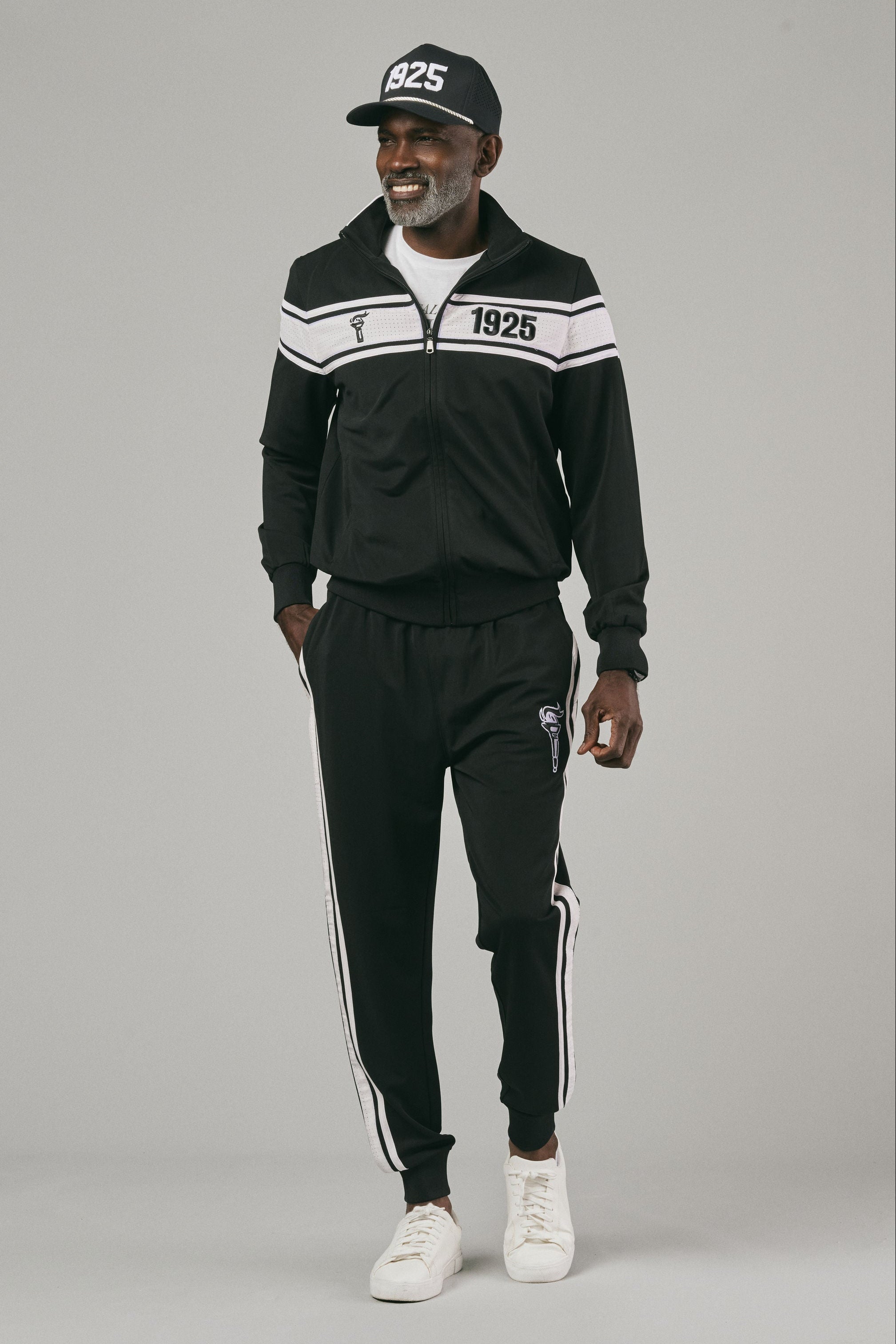 Man wearing a black tracksuit with white stripes and a cap posing against a grey background