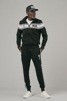 Man wearing a black tracksuit with white stripes and logo and a cap looking sideways a gray background