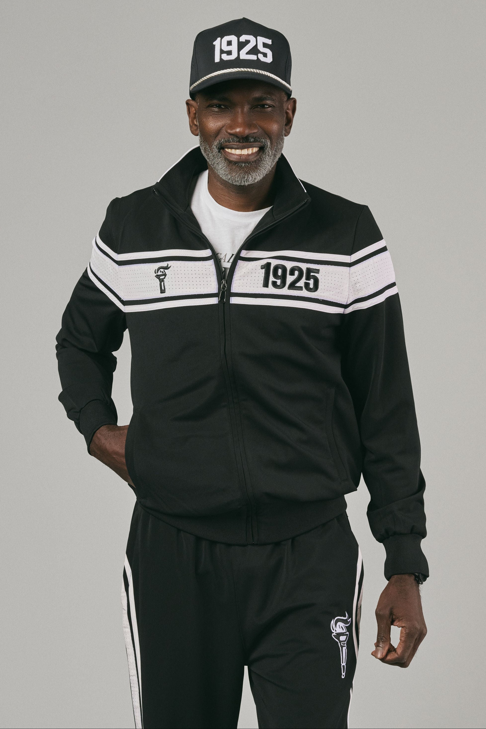 Man wearing a black track jacket with '1925' branding and logo, and black track pants on a grey background