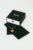 Gold necklace with a pendant on a black card inside an open black box labeled 'The Bar'.